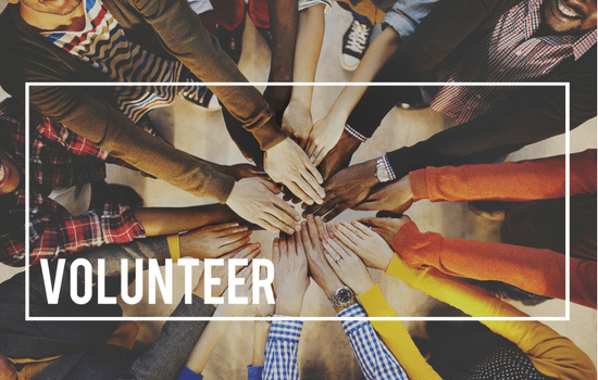 Volunteers standing in a huddle with their hands in. The word VOLUNTEER is in the lower left-hand corner.