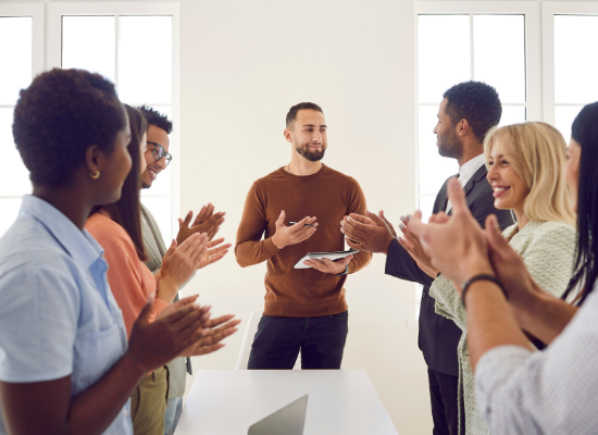 A group of employees are standing around a table congratulating a member of their team with applause.