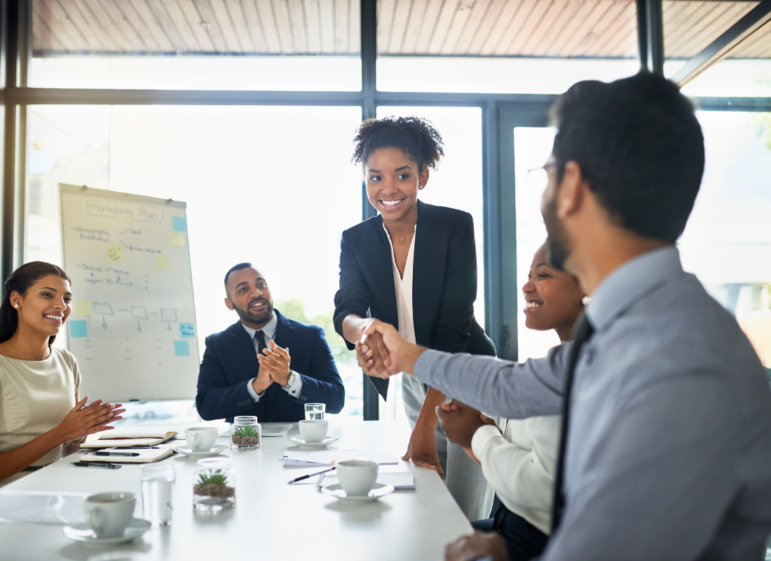 People dressed in business attire are in an office room sitting at a table. Two of them are shaking hands and the rest are applauding.