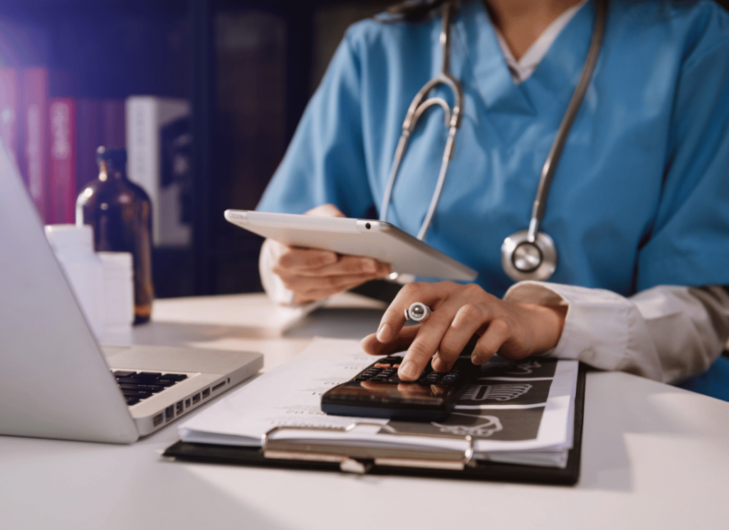 A physician is sitting at a desk reviewing finances for her independently owned physician practice on a tablet and laptop computer while using a calculator.