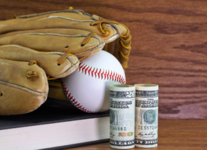 A text book, baseball, leather glove, and dollars are shown on a wood background, symbolizing student athlete compensation.