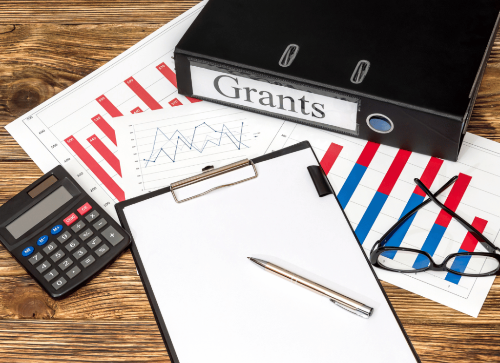A calculator, clip board, eye glasses, and printed graphs are next to a large folder that says "Grants" displayed on a wood background .