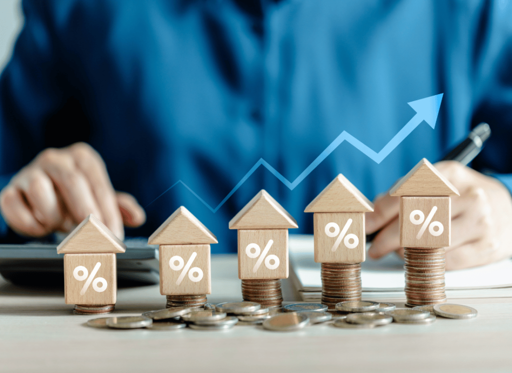 Wooden house models with percentage symbols sit on stacks of coins, representing real estate investment growth. In the background, a real estate investor is assessing the increasing value of the real estate investments.