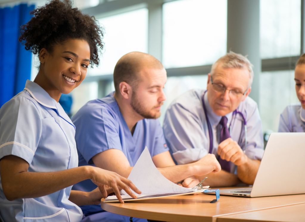 Medical practice staff in a meeting, with one smiling nurse holding paperwork while others review information on a laptop.