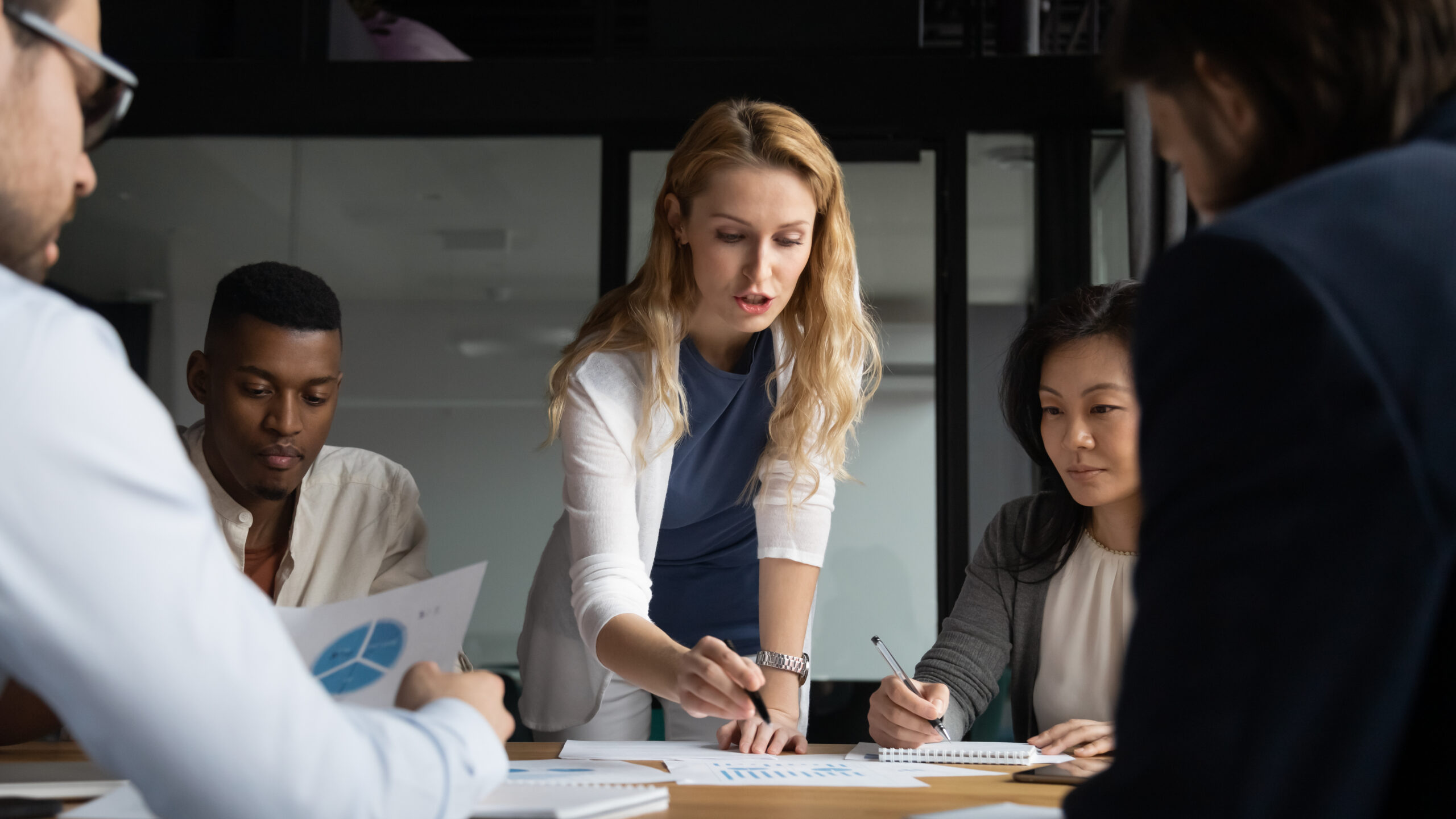 Concentrated young businesswoman explaining market research results in graphs to mixed race colleagues. Focused group of diverse employees holding brainstorming meeting, discussing project ideas.