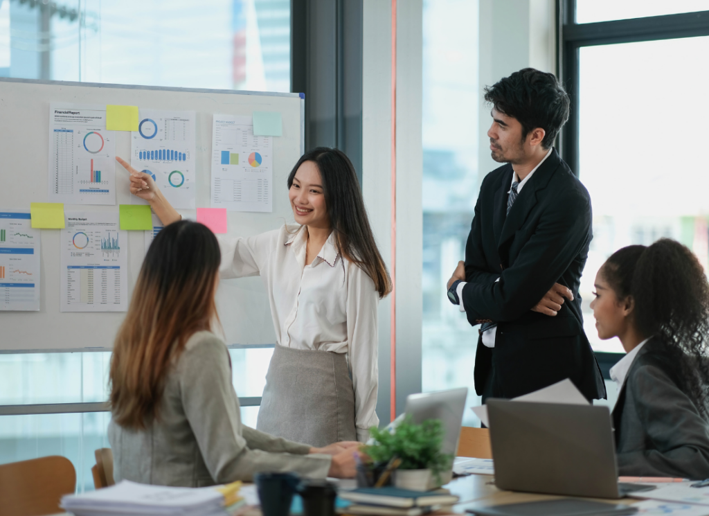 A bookkeeping manager at a nonprofit organization is addressing her team in a board room and pointing to financial reports on a white board.