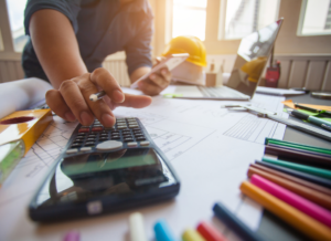 A construction professional calculates tax using a calculator while reviewing blueprints and paperwork, surrounded by office supplies and a hard hat.