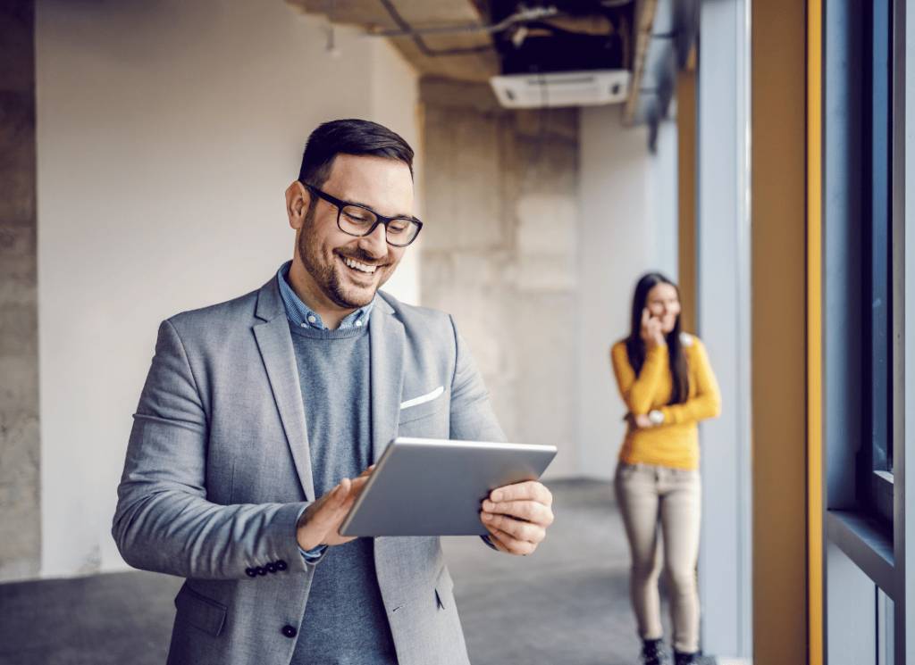 Smiling real estate investor is using a tablet, illustrating strategic long-term maintenance planning for property investments. A colleague stand in the background on the phone.