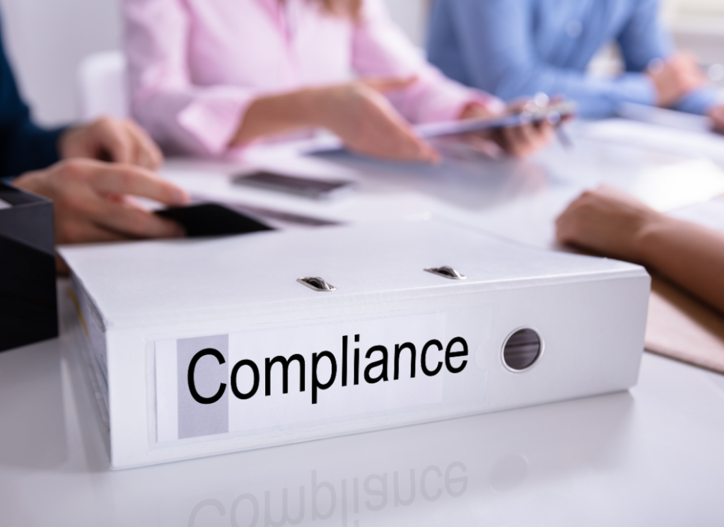 A close-up of a binder labeled "Compliance" on a desk, with people in the background reviewing documents, representing regulatory and compliance requirements for 501(c)(3) organizations.