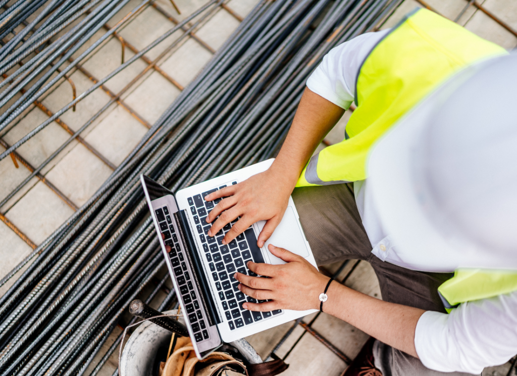 A construction worker in a hard hat and reflective vest uses a laptop while sitting on rebar at a job site seeking information about construction tax credits.