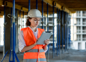 A construction worker wearing a hard hat and orange safety vest reviews information regarding about construction tax planning strategies on a tablet at a construction site.