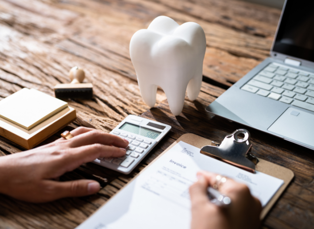 A person uses a calculator while writing on an invoice clipboard at a desk, with a model of a tooth and a laptop nearby. The image represents dental practice bookkeeping and financial management.