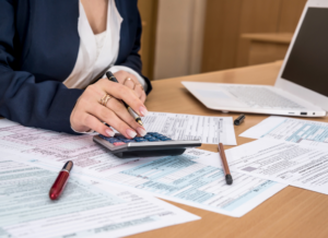 A person in business attire is working at a desk, using a calculator to review and fill out tax forms. Documents and a laptop are spread across the workspace, emphasizing a focus on financial calculations or tax-related tasks.