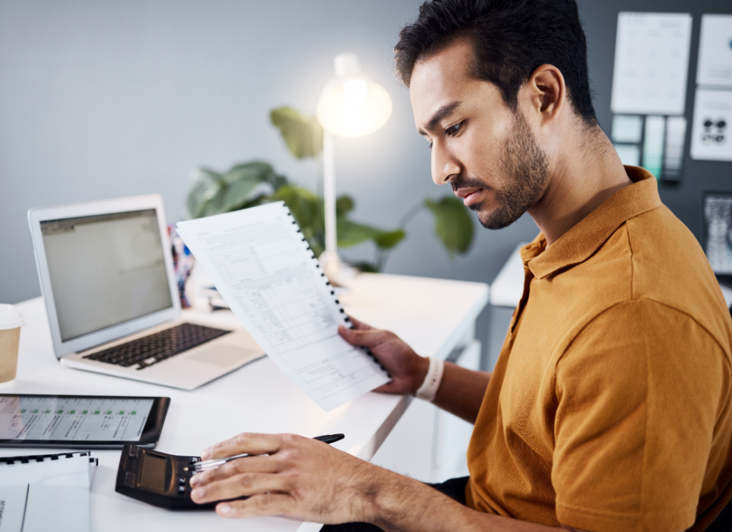 An accountant at a higher education institution sitting at a desk, reviewing documents regarding the use of tax-exempt bonds. The accountant is using a calculator, with a laptop and tablet in the background.