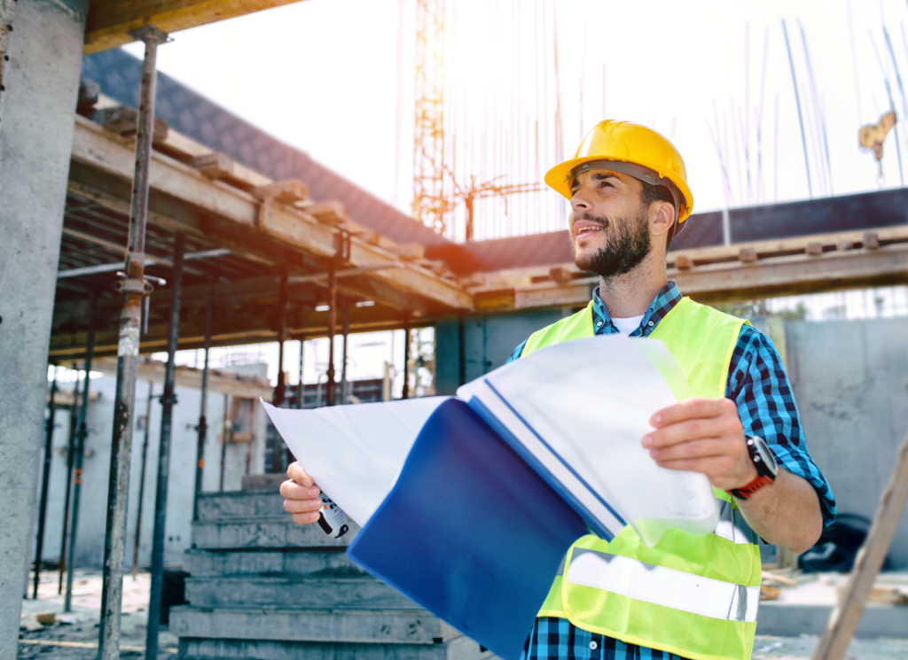 A construction worker wearing a hard hat and reflective vest reviews blueprints at an active construction site, symbolizing project management and planning.