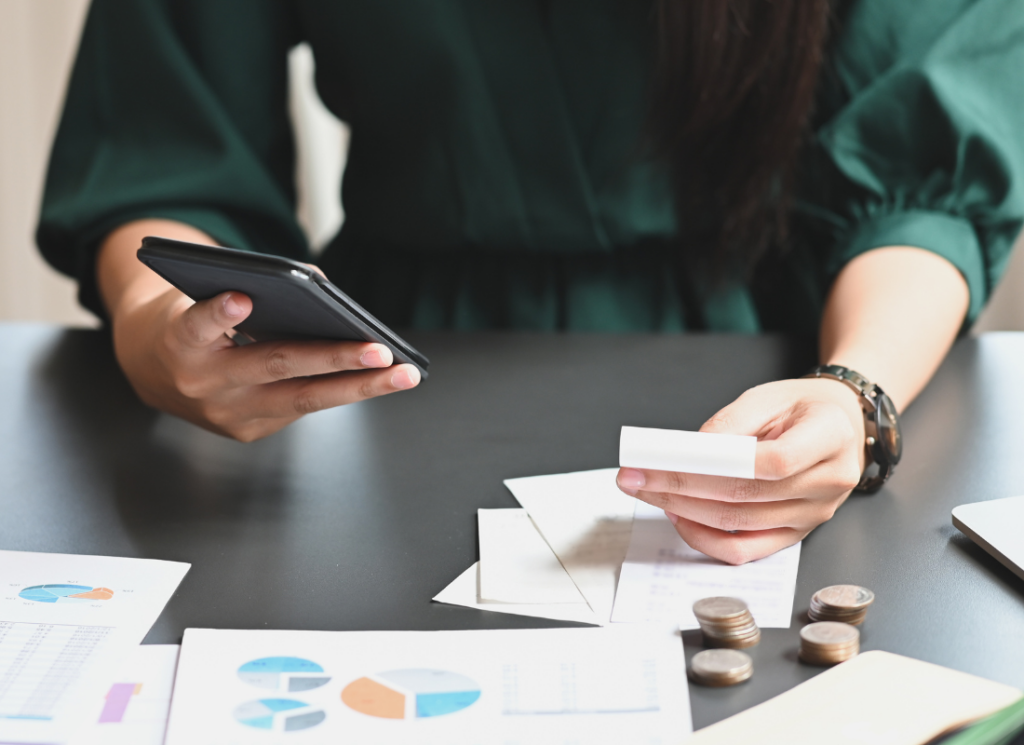 A person holds a smartphone in one hand and a receipt in the other, with financial charts and coins spread on the table, suggesting the process of reviewing expenses or analyzing financial data.