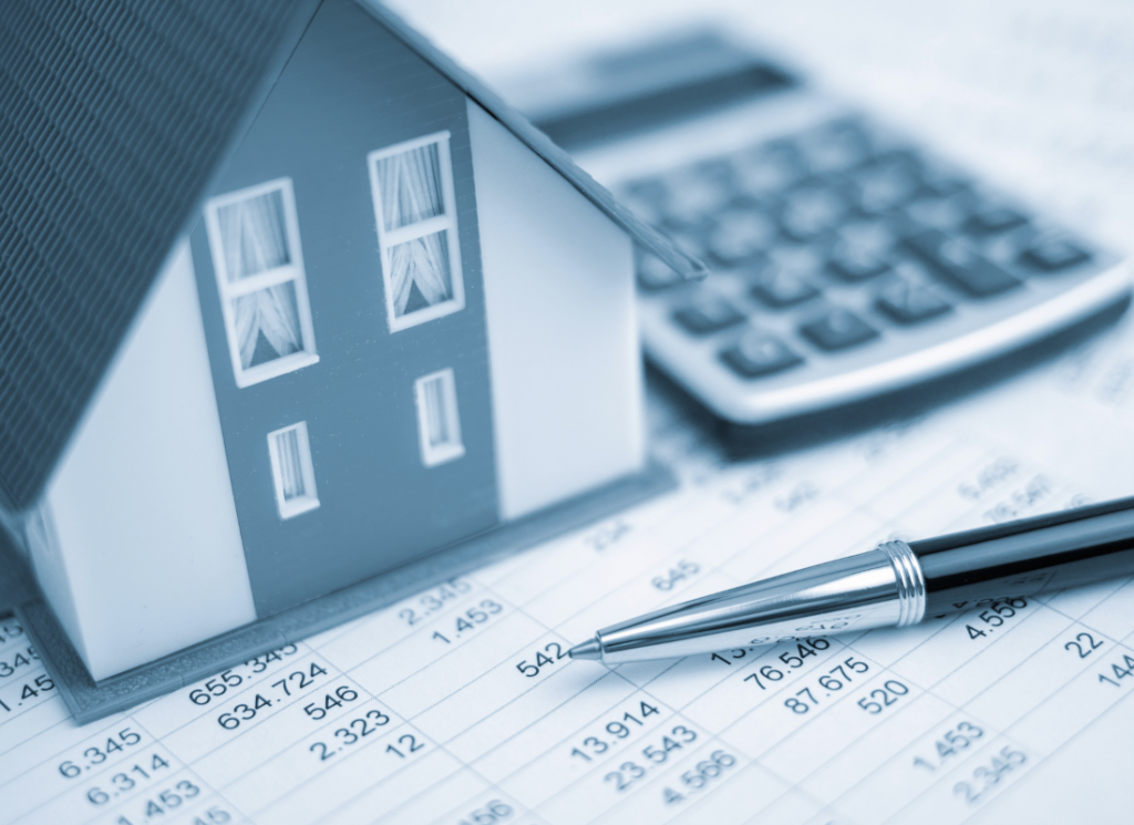 A small model of a house sits on financial documents next to a pen and a calculator, symbolizing real estate investment and financial planning.