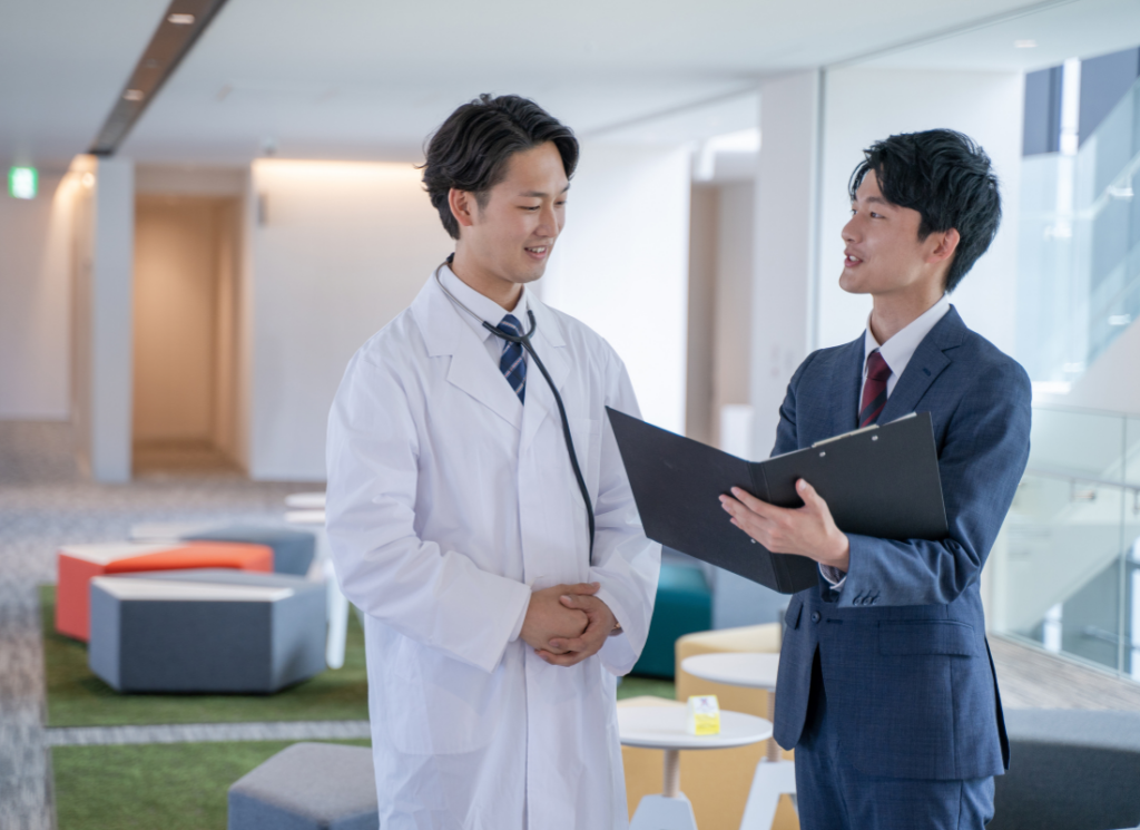 A physician in a white coat and a business professional holding a clipboard are in a discussion about medical practice business structure and financial management.