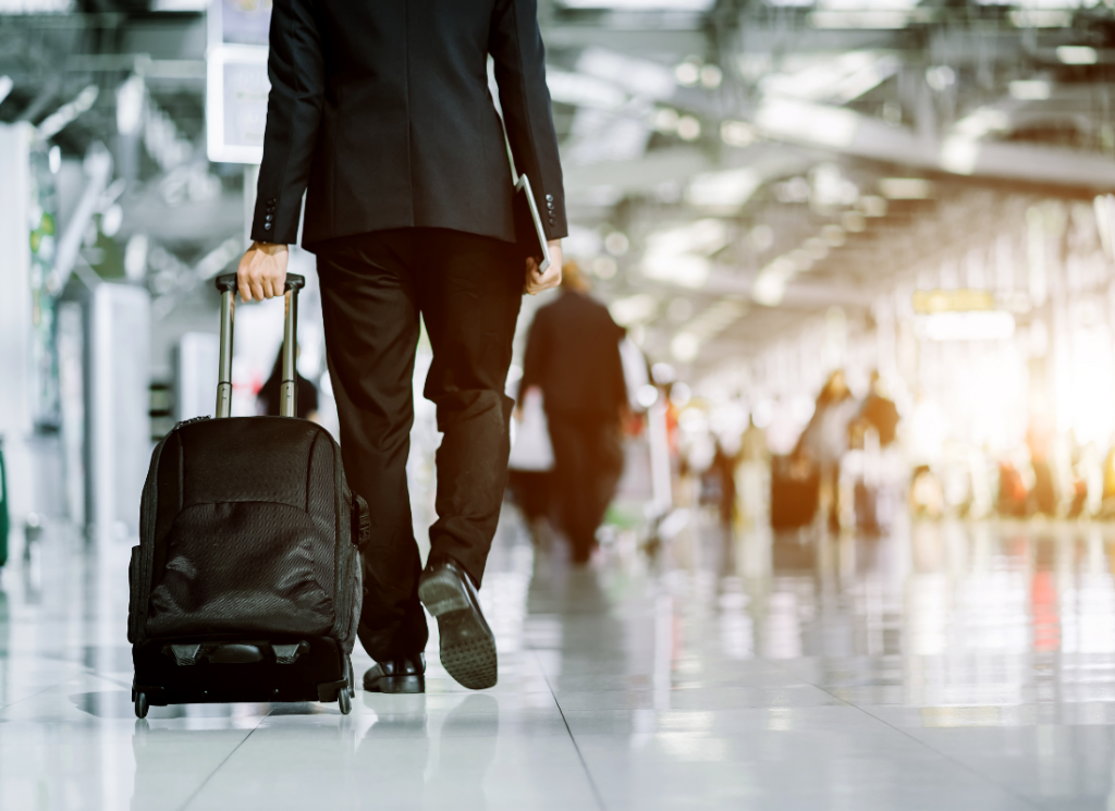 A businessperson in a suit walks through an airport with a rolling suitcase and a laptop, suggesting travel for business purposes.