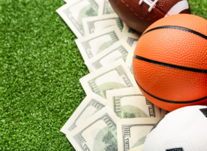 A close-up of a football, basketball, and soccer ball resting on a grassy surface, with several hundred-dollar bills underneath, symbolizing the budget allocated to each of sport in collegiate athletics finance.