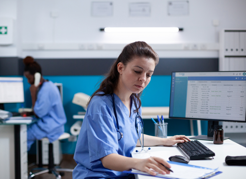 A nurse in scrubs reviews paperwork at a desk while another healthcare professional works in the background, highlighting employees in a physicians office environment.