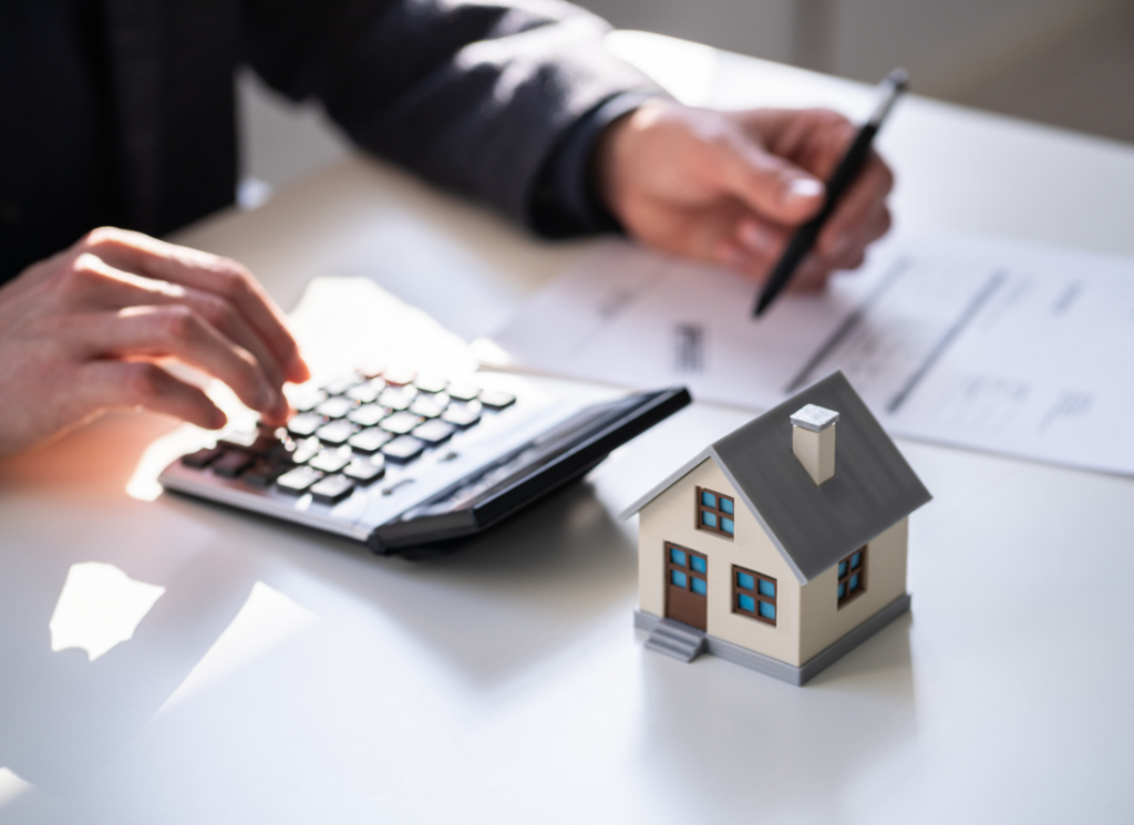 An accountant is sitting at a desk next to a small model house and using a calculator to make financial calculations related to home sale gain exclusion.