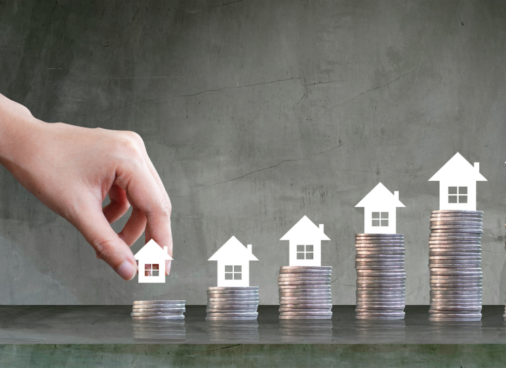 A hand places a house icon on a stack of coins, symbolizing the growth of real estate investments.