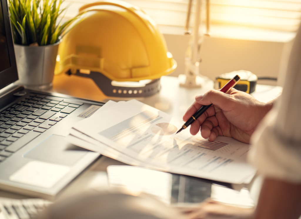 A construction professional reviews financial documents at a desk with a laptop and a hard hat, representing revenue recognition in construction.