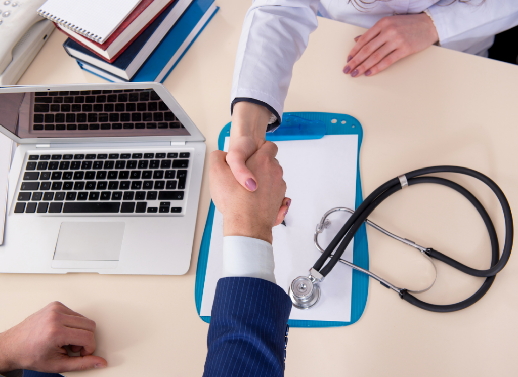 A handshake between a healthcare professional and a healthcare CPA at a desk with a stethoscope, clipboard, and laptop, symbolizing collaboration with a healthcare accounting team.