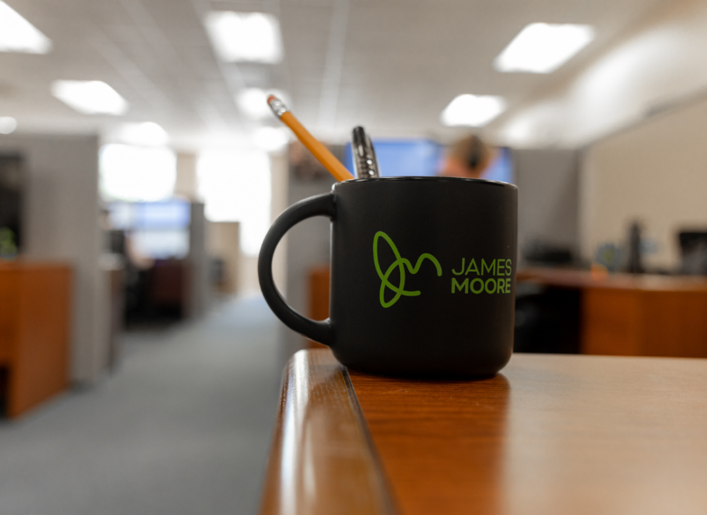 A black James Moore-branded coffee mug filled with office supplies sits on a desk in a professional workspace, representing the firm's office culture and commitment to excellence.