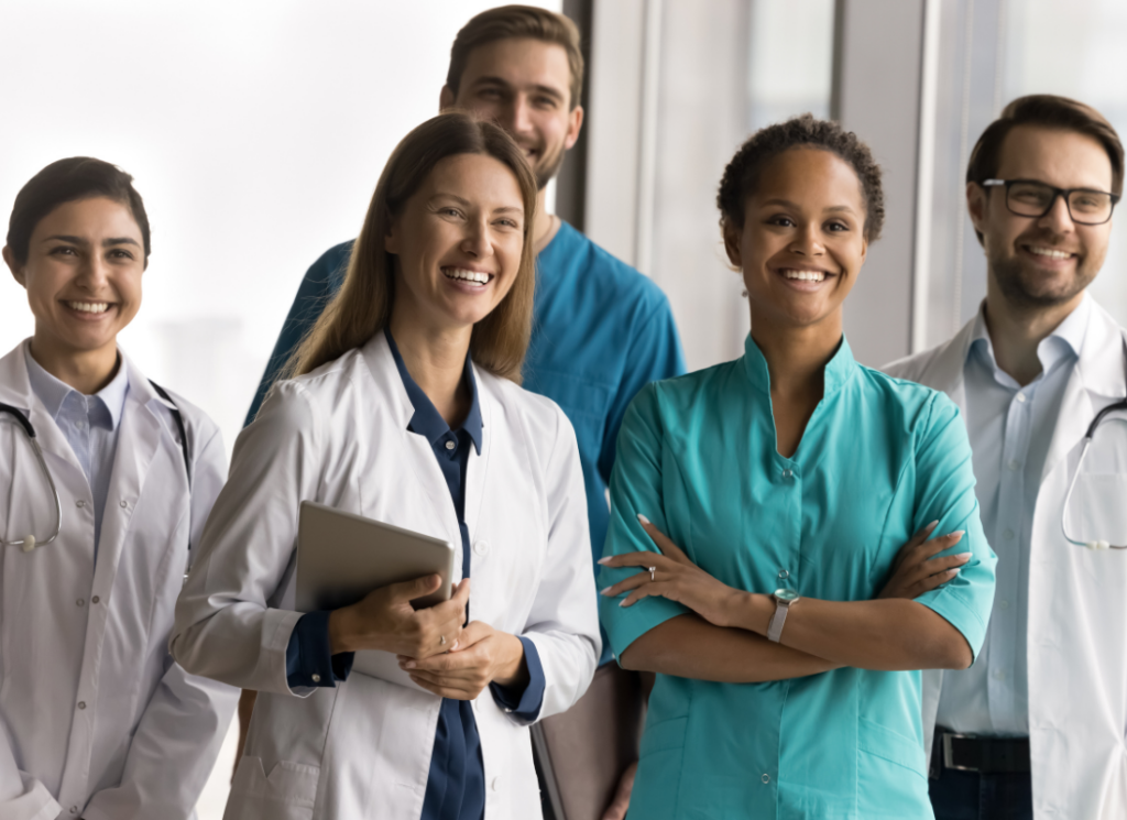 A diverse group of smiling healthcare professionals, including doctors and nurses, stand together in a bright medical facility, representing teamwork and patient care.