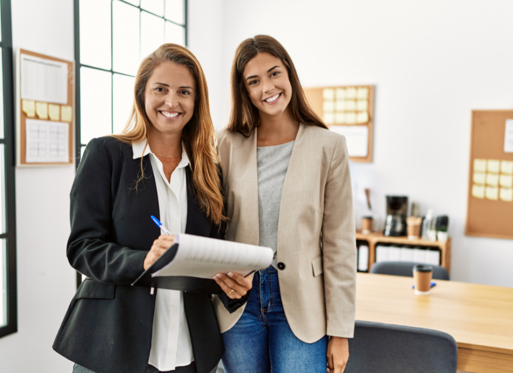 Two professionally dressed women smiling in an office setting, holding documents and collaborating on real estate investment strategies for family offices.