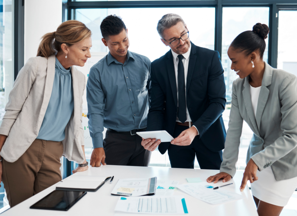 Four professionals collaborate in a modern office, reviewing financial documents and data on a tablet. The image represents teamwork and continuity in government finance.