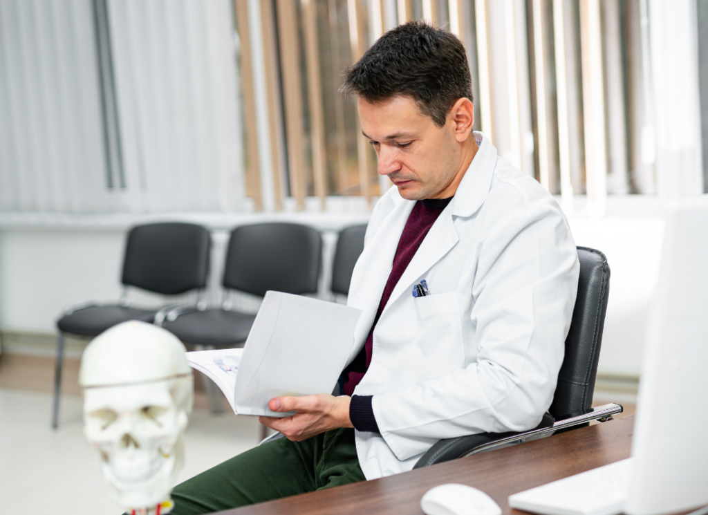 A physician in a white coat sits at a desk, reviewing documents in a medical office. A skull model and computer are nearby, symbolizing the setup of a new practice in Florida.