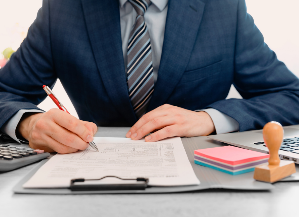 A professional in a suit reviews and signs a financial document on a clipboard, with a calculator, laptop, and office supplies nearby, symbolizing outsourced finance in local government.