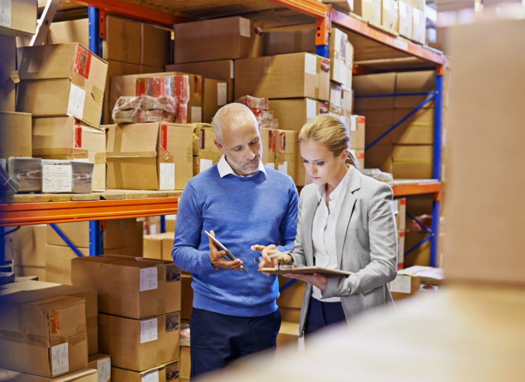 Two professionals stand in a warehouse filled with stacked boxes, reviewing documents on clipboards—discussing inventory, logistics, or transition planning.