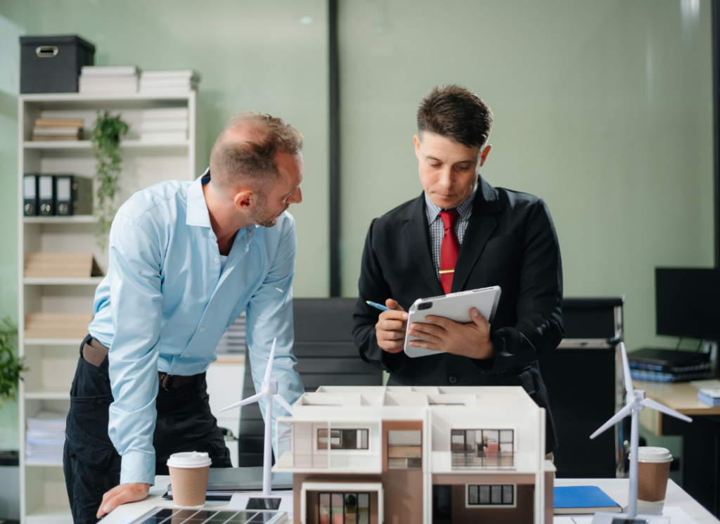 A real estate developer and real estate accountant discuss a project in an office. One takes notes on a tablet while the other leans on a table where architectural models and wind turbines are displayed.